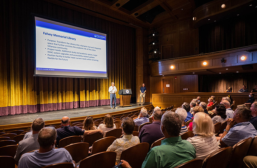 two men stand on stage before a seated audience at the Topper Theatre with a screen in the background displaying bullet points about the Falvey Memorial Library