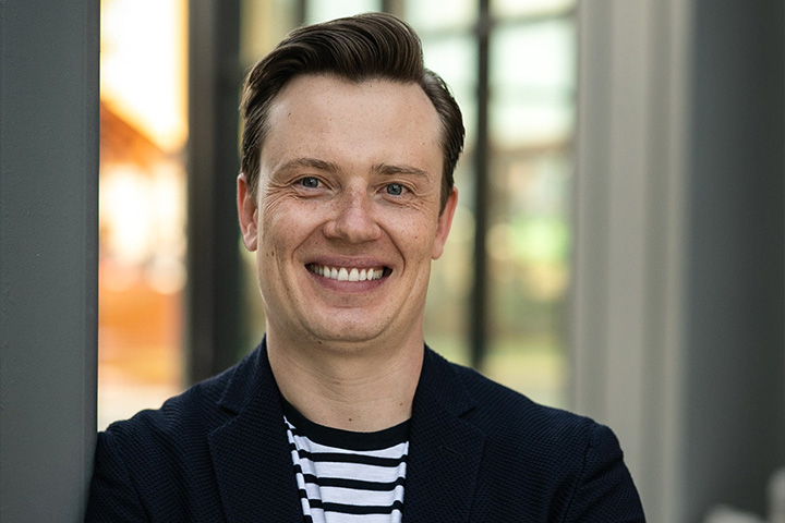 Environmental headshot of Dieter Bender smiling directly at the camera wearing a black suit jacket and shirt with horizontal stripes. 