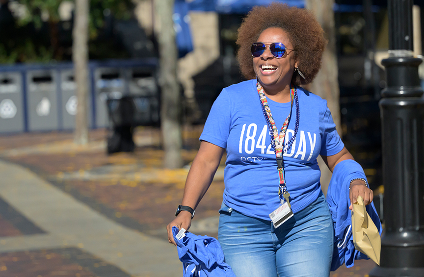 A woman wearing sunglasses and a blue 1842 T-shirt smiles while holding more T-shirts as she walks outside.